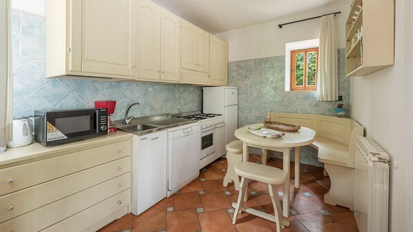 Cocina compacta con armarios blancos, estufa a gas, microondas y una pequeña mesa de comedor con una silla.