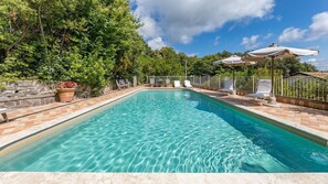 Una piscina rectangular con agua azul cristalina, rodeada por un solárium con baldosas y una valla metálica. Hay tumbonas y sombrillas a lo largo de la piscina.