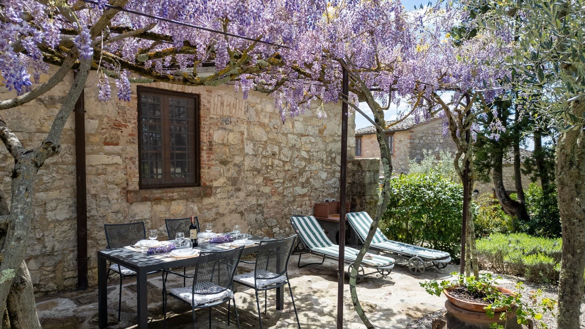 A stone building with a table and chairs, a window, and a pergola with purple flowers.