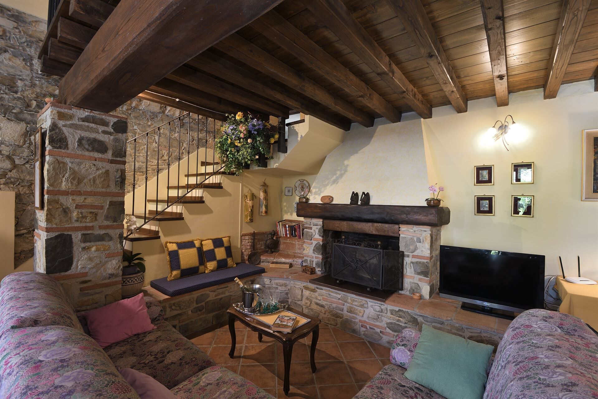 A cozy living room with a stone fireplace, wooden beams, a stone staircase, and a flat-screen TV.