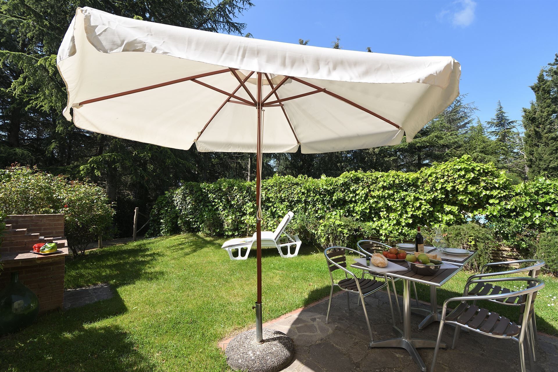 An outdoor patio area with a table and chairs, a white umbrella, and a garden backdrop.