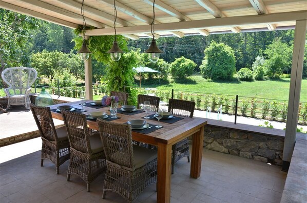 Un patio al aire libre con una mesa de madera dispuesta para comer, sillas de mimbre, luces colgantes y vistas a un jardín exuberante.