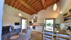 Amplia sala de estar con chimenea, sillones a rayas, una mesa de comedor y una cocina con armarios blancos y salpicadero de piedra.