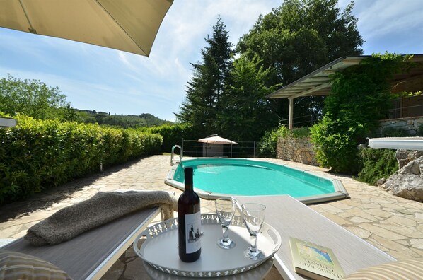 Escena junto a la piscina con una silla de descanso, una botella de vino y copas sobre una bandeja, rodeado de exuberante follaje y un muro de piedra.