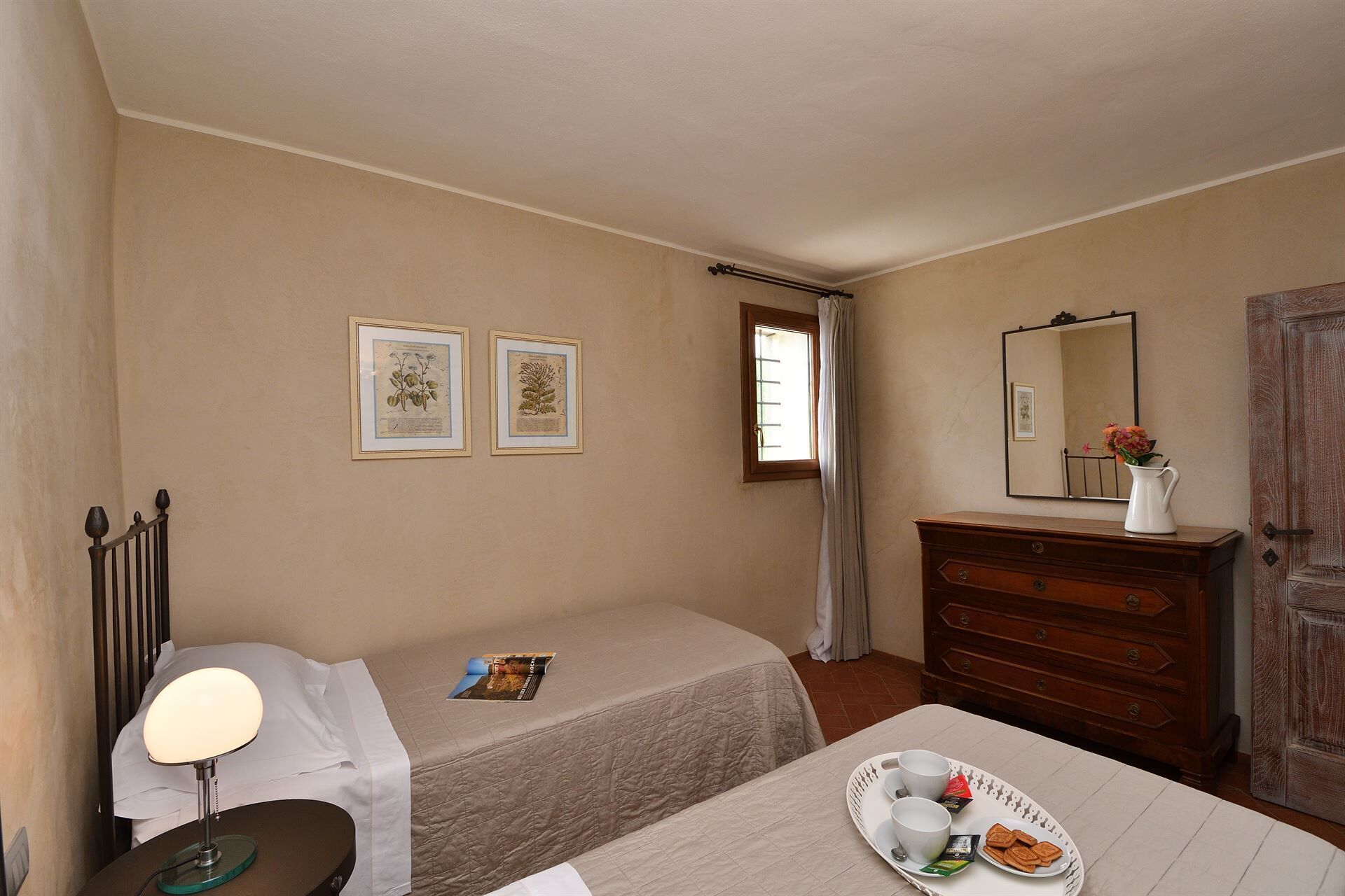 A hotel room with a bed, a bedside table with a lamp, a dresser, a mirror, and two framed botanical prints on the wall.