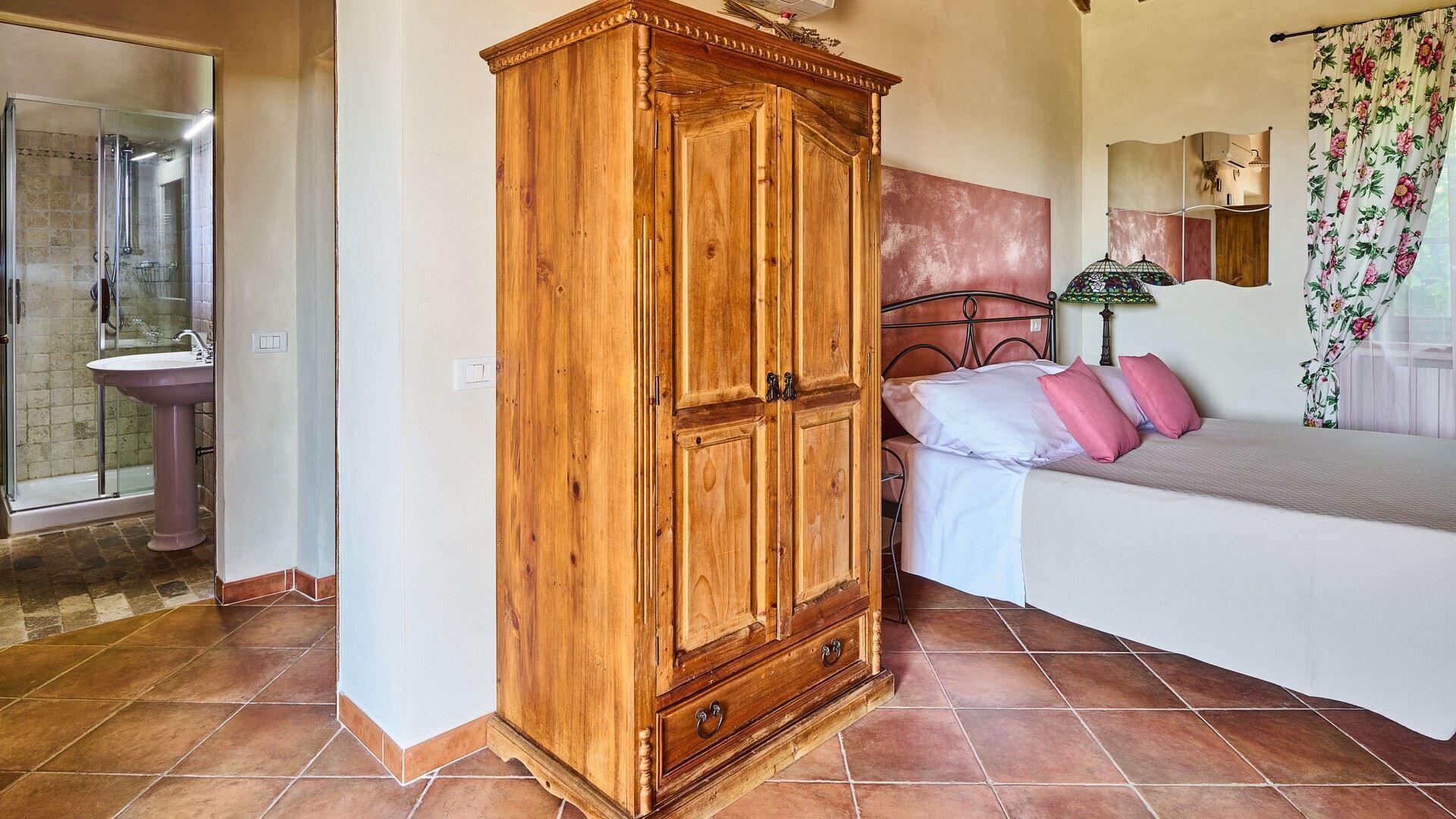 A bedroom with a wooden wardrobe, a bed with pink pillows, a bathroom with a sink, and a shower.