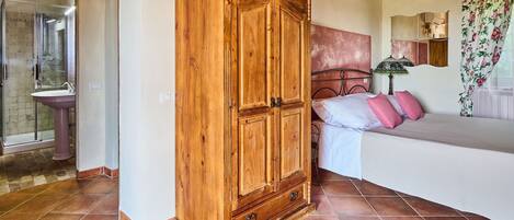 A bedroom with a wooden wardrobe, a bed with pink pillows, a bathroom with a sink, and a shower.