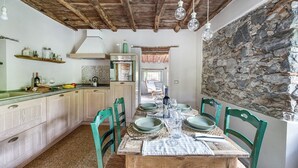 Uma cozinha rústica com armários de madeira, parede de pedra e área de refeições com cadeiras verdes e mesa de madeira posta para dois.
