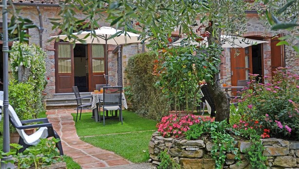 Um pátio com muro de pedra, uma mesa e cadeiras, e uma pequena área de jardim com flores.