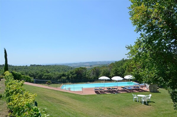 Uma piscina com espreguiçadeiras e guarda-sóis, cercada por gramado e árvores, com vista para a paisagem ao fundo.