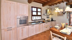 A kitchen with wooden cabinets, a double oven, a window with a cross pattern, a sink, and a dining area with a table set for two. - Ginestra, La Macchia, Peccioli, Pisa and Valdera (Peccioli)