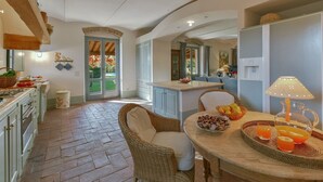 A spacious kitchen with a dining area, featuring a large island with fruit and drinks, a wicker chair, and a view of the garden through the open door.