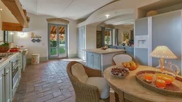 A spacious kitchen with a dining area, featuring a large island with fruit and drinks, a wicker chair, and a view of the garden through the open door.