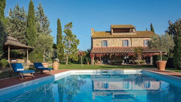 A poolside view of a stone house with a tiled roof, a covered patio, and a garden with trees and a pergola.