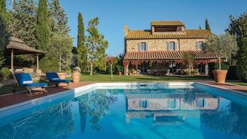 A poolside view of a stone house with a tiled roof, a covered patio, and a garden with trees and a pergola.
