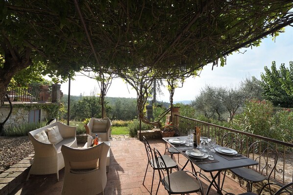An outdoor patio with wicker furniture and a table set for dining, surrounded by trees and a view of the countryside.