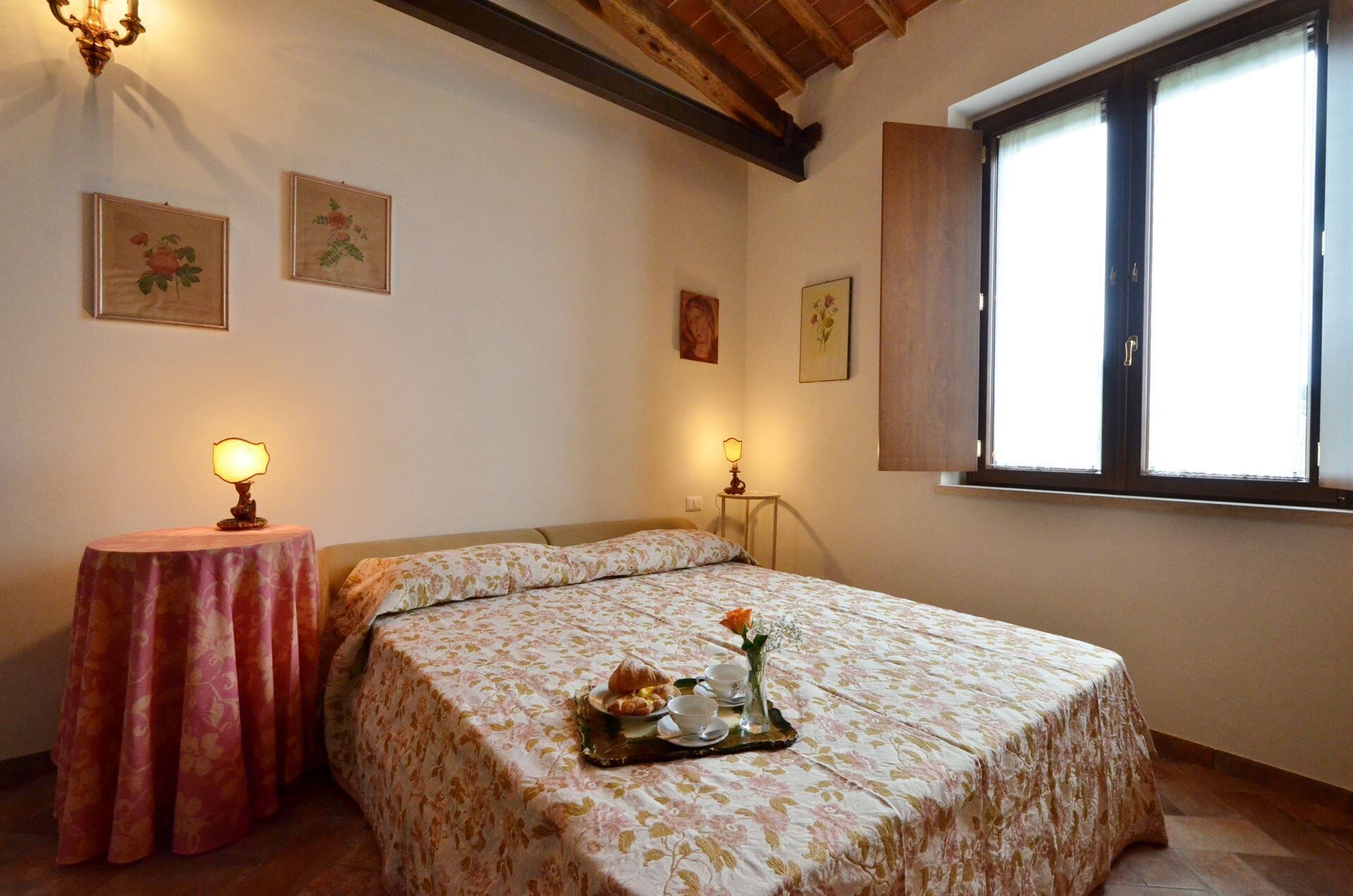 A bedroom with a bed, a bedside table with a lamp, a window with curtains, and floral artwork on the walls.
