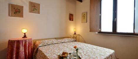 A bedroom with a bed, a bedside table with a lamp, a window with curtains, and floral artwork on the walls.