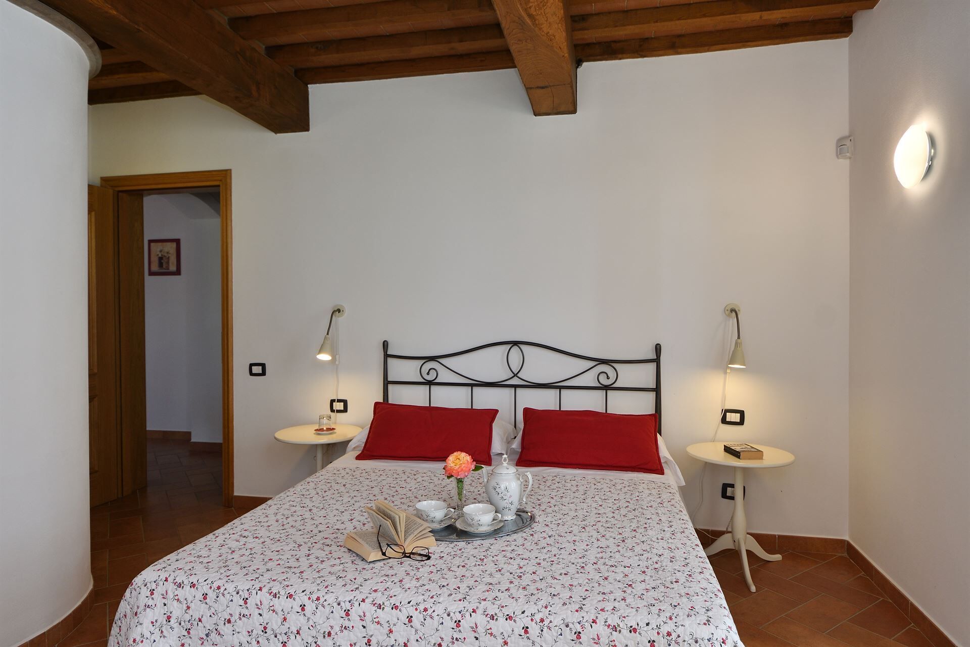 A bedroom with a bed, two bedside tables, a nightstand, a red pillow, a white wall, and a wooden ceiling.