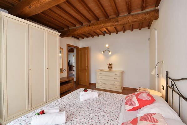 A bedroom with a wooden ceiling, a bed with floral bedding, a dresser, and a nightstand with a lamp. - Papaveri, La Macchia, Peccioli, Pisa and Valdera (Peccioli)