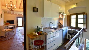 Une cuisine moderne avec des meubles blancs, un évier en acier inoxydable et un plan de travail en bois. Un lustre est suspendu au-dessus de l’évier, et une fenêtre offre une vue sur la verdure à l’extérieur.