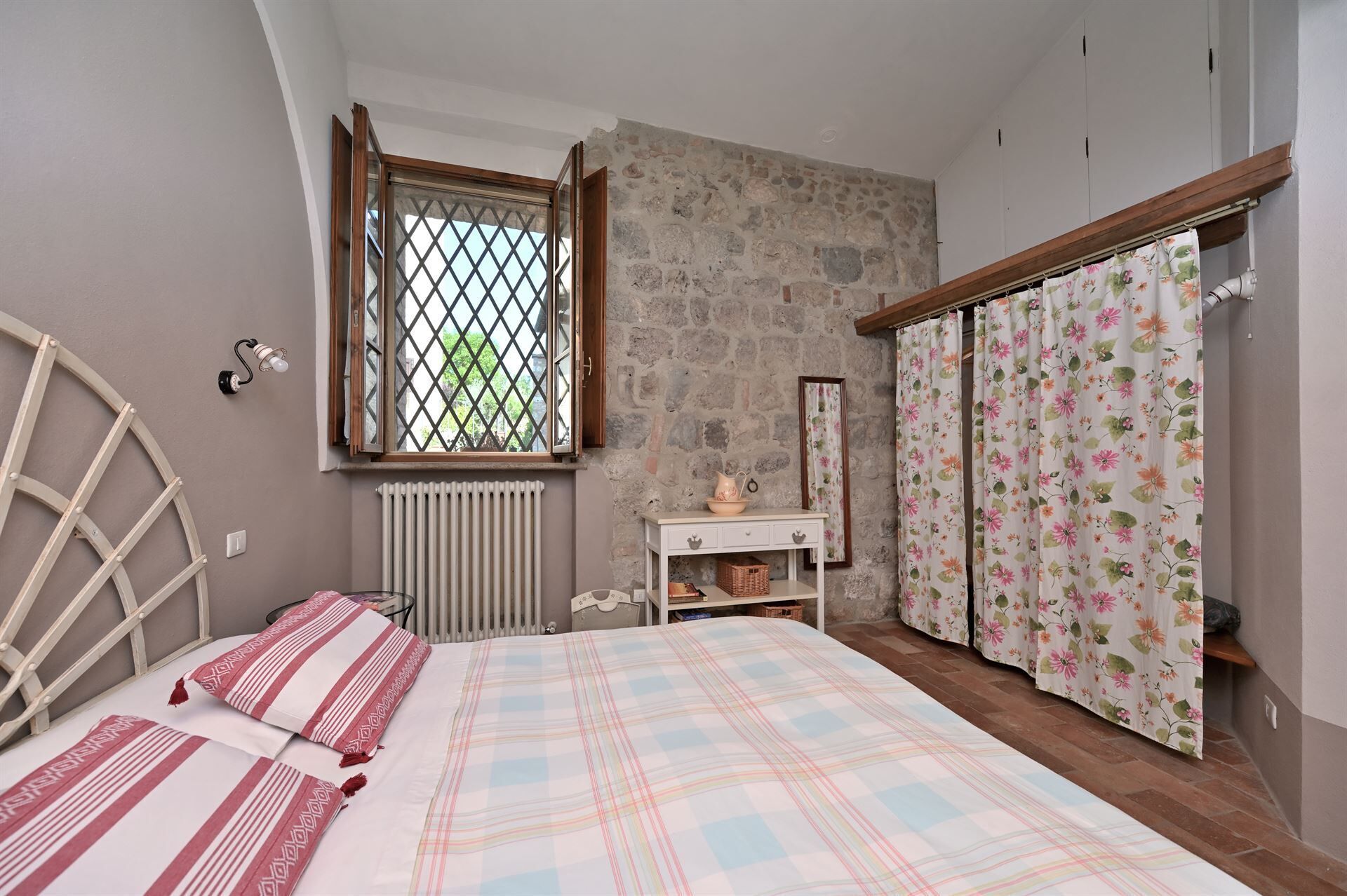 A bedroom with a bed, a radiator, a window with a lattice, a wooden wardrobe, and a floral curtain.