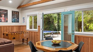 A cozy room with a foosball table, a dining table with a bowl of fruit, and a view of the outdoors through a door. - Getaway in the Forest, Surrounded by Peaceful Nature and Year-round Creek (Cazadero)