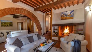 A cozy living room with a fireplace, white sofas, and a coffee table with fruits and wine.