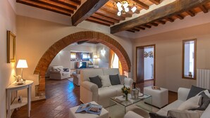 A spacious living area with a fireplace, white sofas, a glass coffee table, and a kitchen in the background.