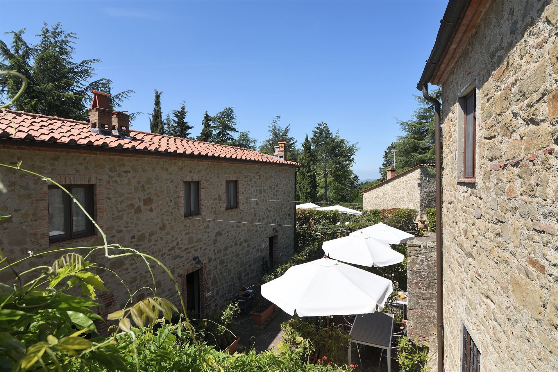 A stone building with a tiled roof, outdoor seating with white umbrellas, and surrounding greenery.