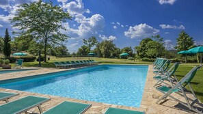 Une piscine rectangulaire à l’eau d’un bleu limpide, entourée de carreaux de pierre. De nombreux fauteuils de jardin et parasols verts sont disposés le long du bord de la piscine.