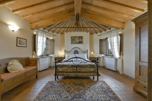 Un dormitorio con techo de madera, una cama con estructura de metal, un banco, un radiador y un ventanal grande con cortinas.