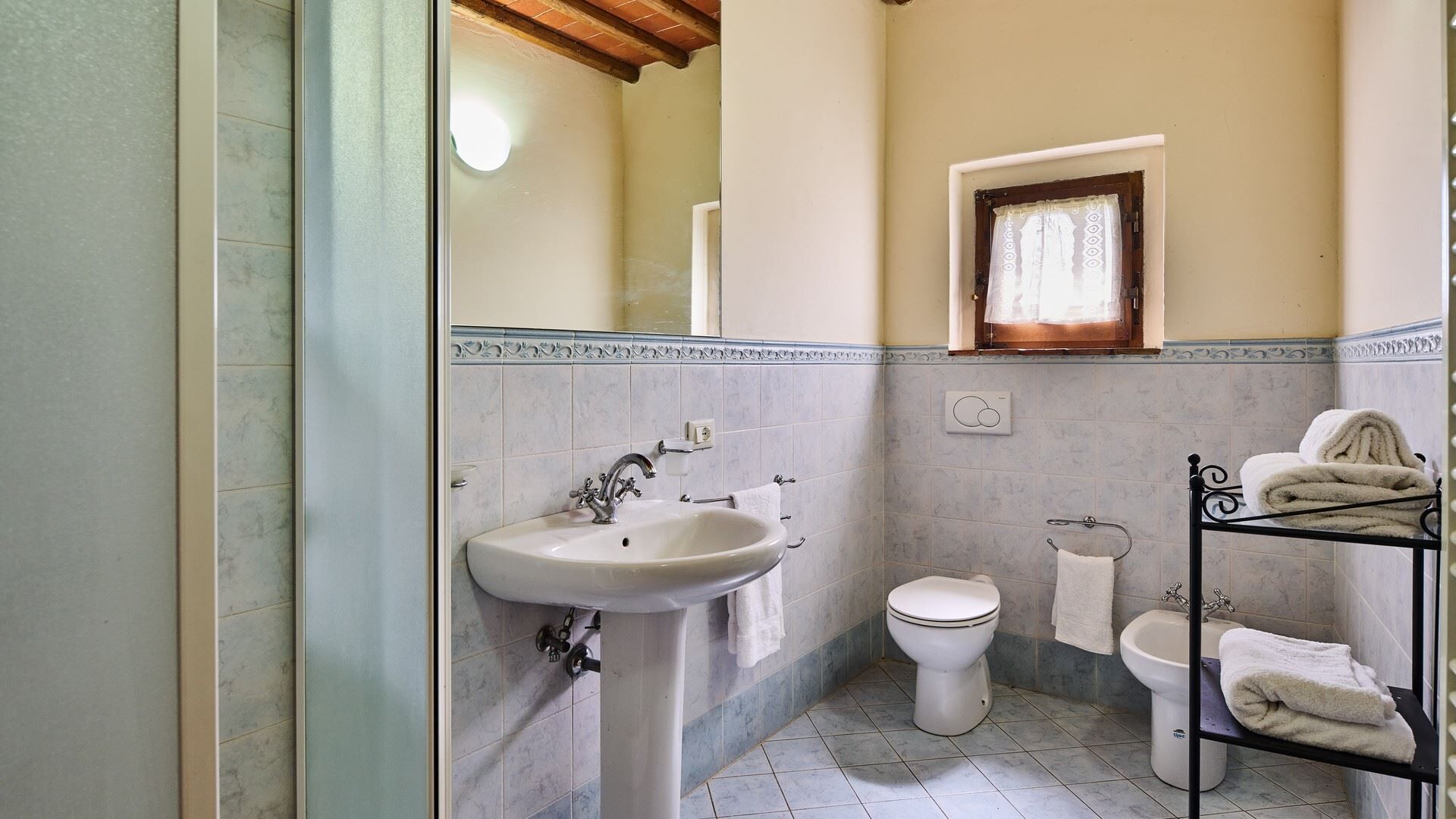 A bathroom with a sink, toilet, and bidet, featuring tiled walls and a window with a curtain.