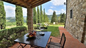 A covered outdoor dining area with a table set for two, surrounded by stone walls and a view of rolling hills and tall trees. - Urania, Podere di Casalta, Castellina in Chianti, Siena and Chianti (Castellina in Chianti)