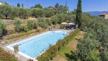 Ein Swimmingpool mit Liegestühlen und Sonnenschirmen, umgeben von viel Grün und einer Steinmauer.