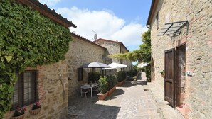 Une ruelle étroite, pavée de pierres, avec une table et des chaises placées sous un auvent, le tout entouré de bâtiments en pierre ornés de lierre et de plantes en pot.