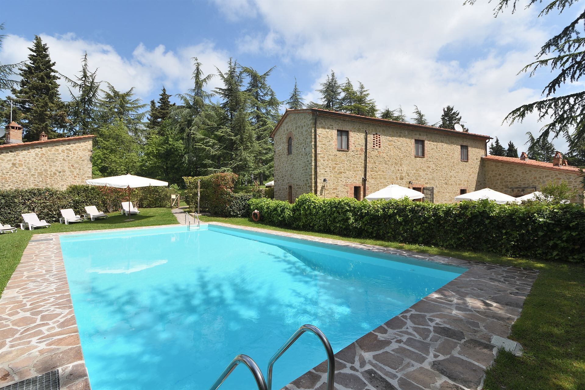 A swimming pool with stone tiles, surrounded by a stone building, a grassy area, and trees.