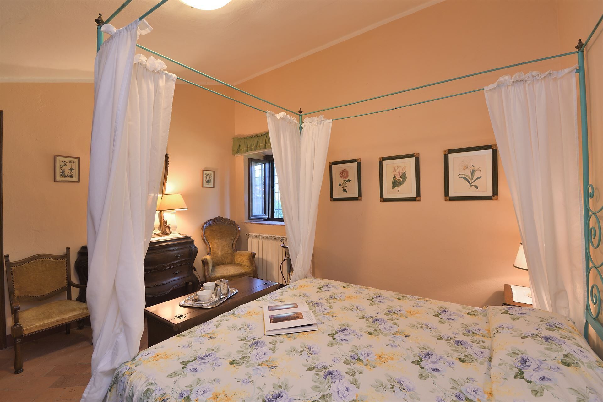 A bedroom with a four-poster bed, a wooden chair, a desk with a lamp, and framed botanical prints on the wall.