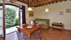 A dining area with a wooden table, chairs, and a green sofa. There is a window with curtains, a wall-mounted light, and a small table with fruits and a bottle.