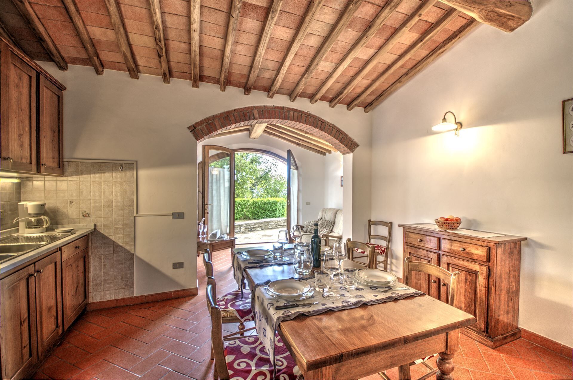 A rustic kitchen with a wooden table set for dining, a coffee machine, and a view through an open archway to a garden.