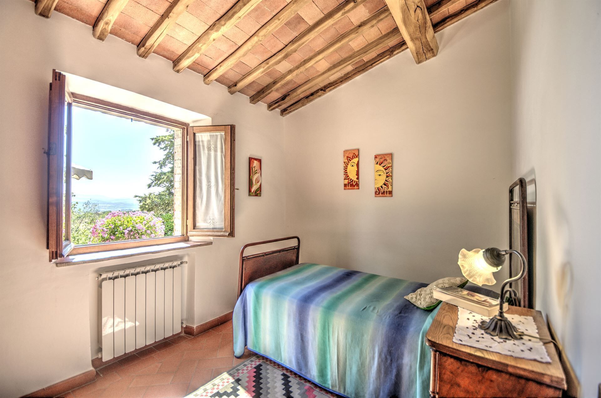 Une chambre avec un plafond en bois, une fenêtre donnant sur la verdure, un lit recouvert d’une couverture colorée, une table de chevet avec une lampe, et deux tableaux accrochés au mur.