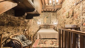 Un dormitorio rústico con pared de piedra, vigas de madera en el techo, una cama con colcha estampada, una silla de mimbre y una mesita con lámpara.