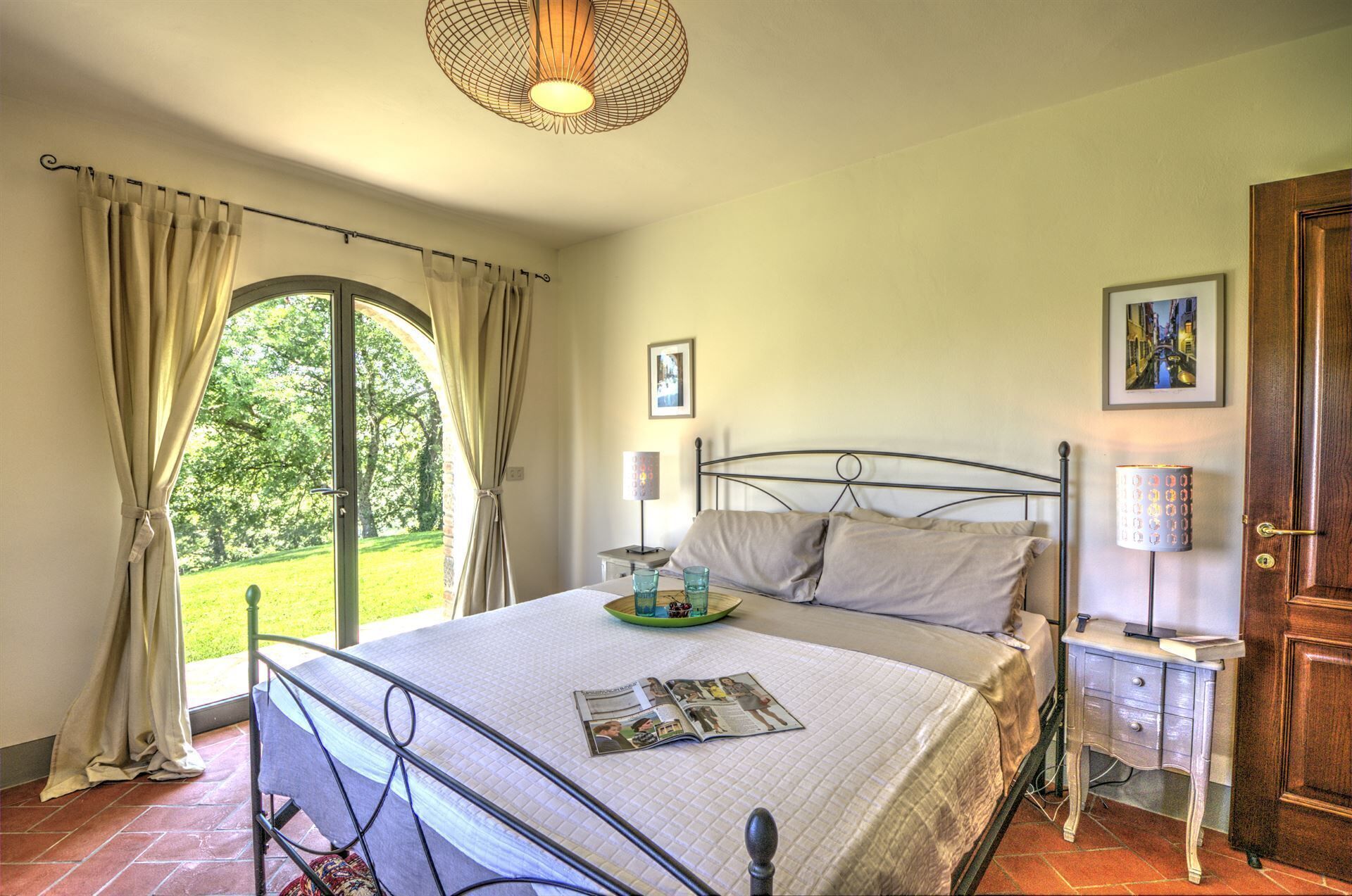 Un dormitorio con cama, mesita de noche, lámpara, un cuadro enmarco y vista a un jardín por la ventana.