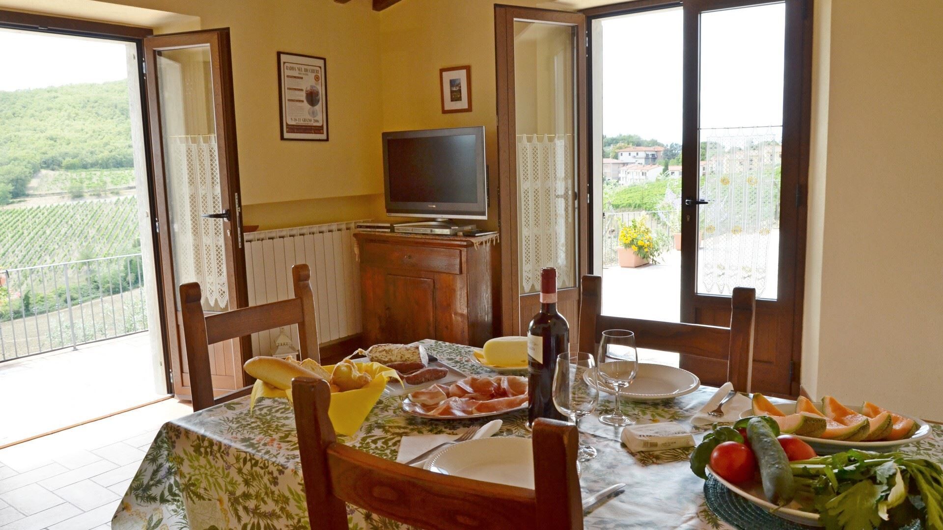 A dining area with a table set for two, featuring a bottle of wine, glasses, plates with food, and a view of a vineyard through open doors.