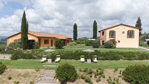 Il s’agit d’une propriété comprenant deux bâtiments principaux, un jardin avec une pelouse et des allées, ainsi qu’un espace extérieur aménagé avec des parasols.