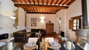 A cozy living room with a wooden ceiling, a fireplace, a sofa, a dining table set with glasses and a bottle, and a stone wall with a framed picture.
