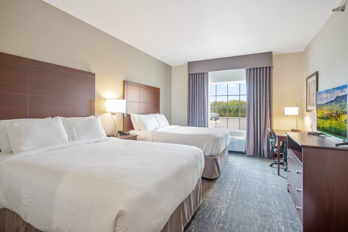 A hotel room with two beds, a desk, a chair, a TV, and a large window with curtains.