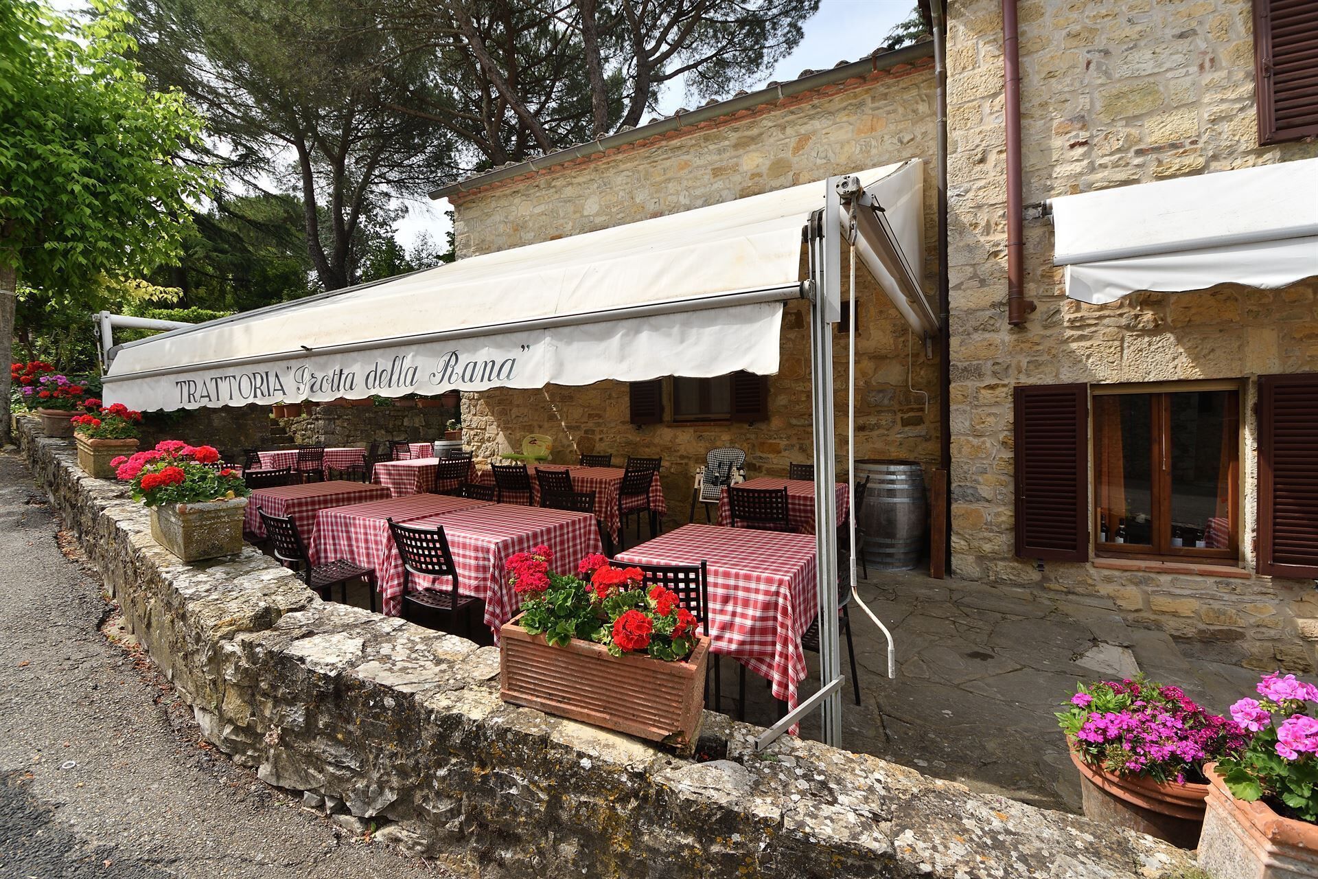 An outdoor seating area with tables covered by red and white checkered tablecloths, surrounded by stone walls and potted plants.