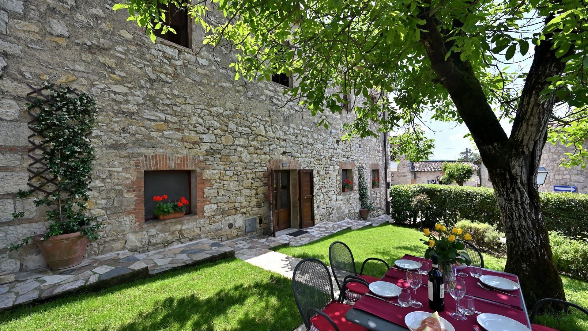Un edificio in pietra con un piccolo patio giardino, un tavolo da pranzo apparecchiato per due e un albero che offre ombra.
