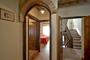 A hallway with a wooden door leading to a room with a red bedspread. A staircase with a wooden railing is visible to the right.